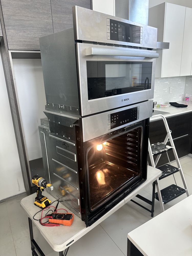 Built-in oven units pulled out of cabinetry, with an open lower oven, multimeter and drill beside them.